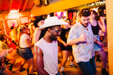 Smiling man in a white cowboy hat and glasses dancing on a lively indoor dance floor with casually dressed people, warm colorful lights, and string-light decor.