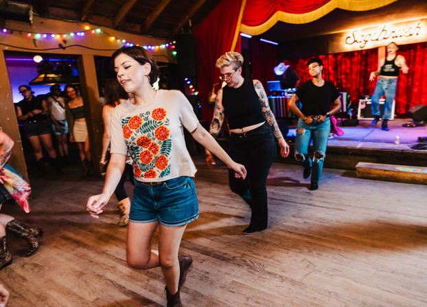 Young adults dancing on a wooden floor in a cozy, neon-lit live music venue; a woman in a floral tee and denim shorts leads the group near a small stage with red curtains and string lights.