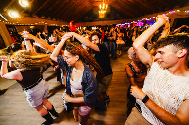 Lively indoor dance party in a wood‑beamed hall — crowded wooden dance floor with people twirling and social dancing under string lights and chandeliers.