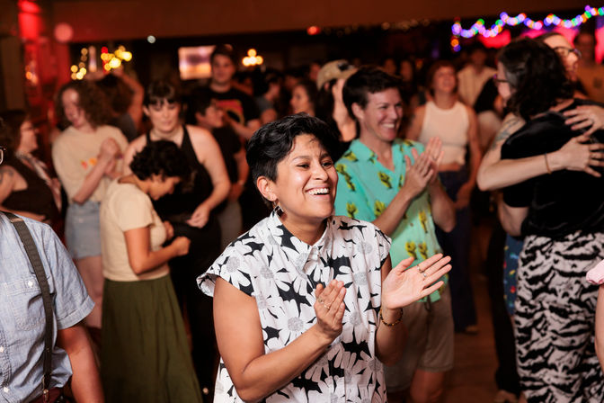 Smiling person in a floral shirt clapping on a lively indoor dance floor with a diverse crowd and string lights