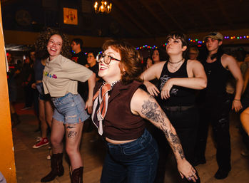 Young adults dancing at a lively indoor music venue — smiling woman with glasses and floral arm tattoos in the foreground, friends in casual and vintage outfits, colorful string lights and wooden ceiling overhead