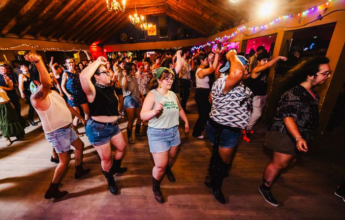 Energetic crowd line-dancing in a rustic indoor dance hall with a wooden vaulted ceiling, string lights and colorful party lighting; people in shorts, boots and casual summer outfits moving in unison.