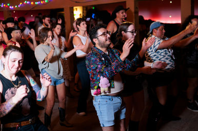 Energetic late-night dance floor in a dimly lit club — a diverse crowd in casual festival outfits clapping and dancing under colorful string lights, including a person in a floral jacket and denim shorts with a small plush toy attached.