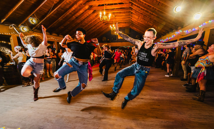 Energetic barn dance scene: joyful crowd line-dancing mid-jump in a rustic wooden dance hall with chandeliers and string lights, dancers in jeans, boots and casual outfits