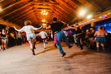 Lively barn-style indoor dance hall with a crowd mid-step line dancing on a wooden floor, vaulted timber ceiling and chandeliers, colorful string lights and spectators around the perimeter.