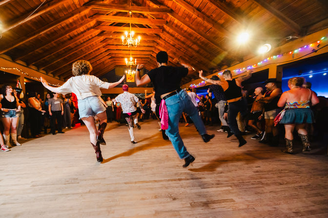 Lively barn-style indoor dance hall with a crowd mid-step line dancing on a wooden floor, vaulted timber ceiling and chandeliers, colorful string lights and spectators around the perimeter.