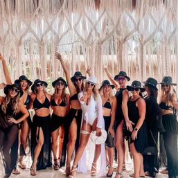 Bride-to-be in a white swimsuit and heart sunglasses posing with bridesmaids in black swimwear and wide-brim hats under a boho macrame fringe canopy at a tropical beach resort