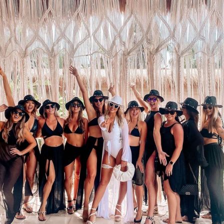 Bride-to-be in a white swimsuit and heart sunglasses posing with bridesmaids in black swimwear and wide-brim hats under a boho macrame fringe canopy at a tropical beach resort