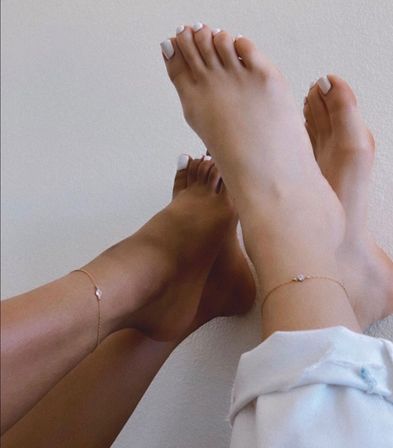 Close-up of two crossed bare feet with white pedicure and delicate gold anklets against a neutral wall — minimalist, relaxed lifestyle shot