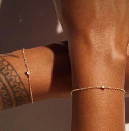 Close-up of two sunlit wrists with warm brown skin wearing delicate gold chain bracelets with tiny diamond-like charms and a geometric wrist tattoo.