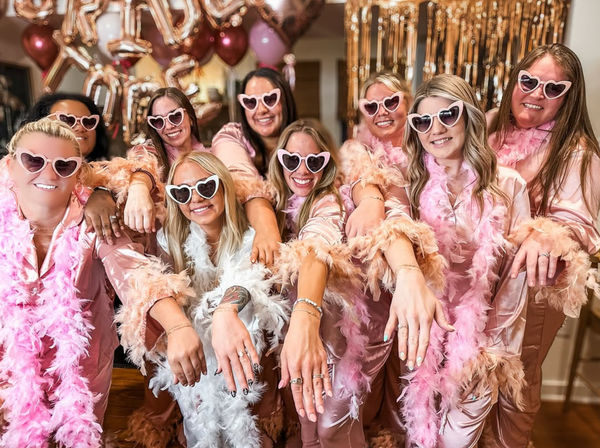 Pink-glam bachelorette crew in matching satin robes, heart-shaped sunglasses and feather boas showing off rings and hands in front of rose-gold balloons and a gold fringe backdrop