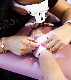 Close-up of a manicurist trimming a client’s cuticles under a white magnifying loupe on a pink salon table, hands wearing gold rings and bracelet