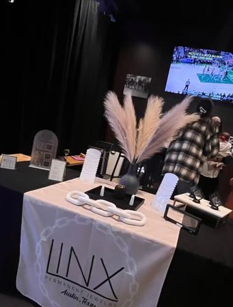 Pop-up jewelry display table with pampas grass centerpiece and decorative white chain at an indoor Austin, Texas event, TV showing basketball in the background.