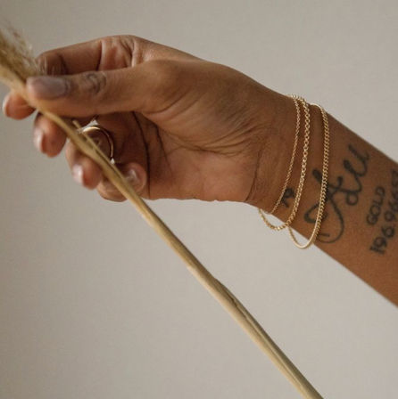 Close-up of a hand wearing delicate gold chain bracelets and a script tattoo, holding a dried pampas grass stem against a soft neutral background.