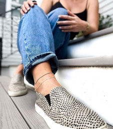 Close-up of a person wearing rolled blue denim jeans and spotted slip-on sneakers with delicate double ankle chains, seated on gray porch steps in a casual summer outfit