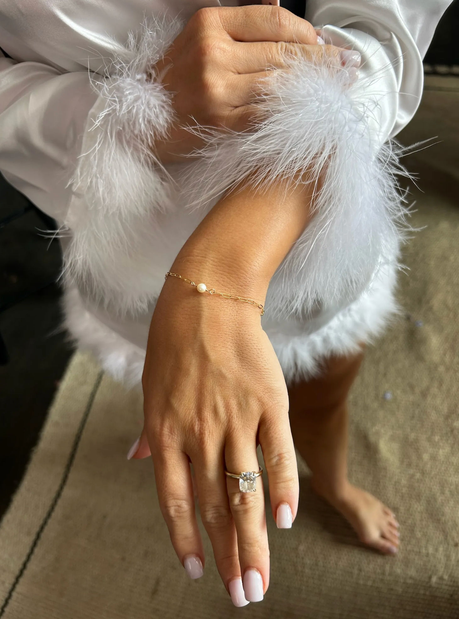 Close-up of hand wearing a cushion-cut engagement ring and delicate gold chain bracelet with a single pearl, pale-pink manicure and white feather-trimmed robe sleeve over a neutral rug
