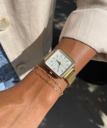 Sunlit close-up of a tan wrist wearing a minimalist square white-dial gold mesh watch and two delicate gold chain bracelets, paired with a rolled white blouse cuff.