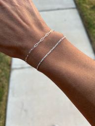 Sunlit close-up of a wrist wearing two delicate silver chain bracelets above a concrete sidewalk and grass