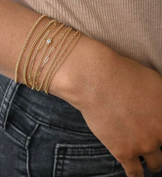 Close-up of a wrist wearing stacked delicate gold bracelets — mixed chains with one small bezel-set gemstone — styled with a beige ribbed top and dark denim jeans.