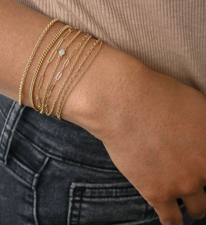 Close-up of a wrist wearing stacked delicate gold bracelets — mixed chains with one small bezel-set gemstone — styled with a beige ribbed top and dark denim jeans.