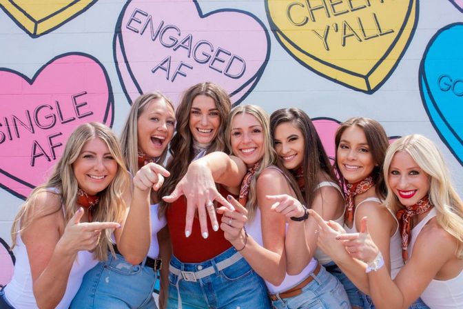 Engagement celebration: seven friends pose in front of a colorful heart-shaped mural as the newly engaged woman shows her ring while everyone points and smiles.