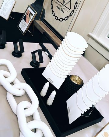 Minimalist pop-up jewelry display with delicate gold chain necklaces on white cards, faceted ring cones, a textured black tray and oversized white decorative chain on a table.