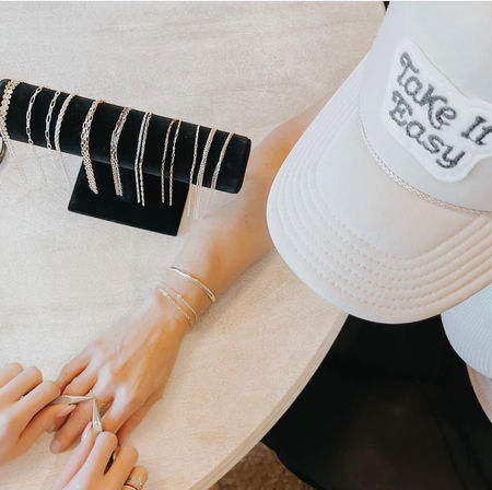 Close-up of hands adjusting a delicate silver ring on a wrist with dainty chain bracelets, black velvet T-bar displaying multiple silver chain necklaces on a light wood table, and a white baseball cap with a "Take it Easy" patch.