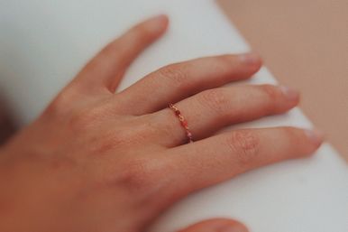 Close-up of a hand resting on a white surface, wearing a delicate gold chain ring dotted with tiny pink and red beads.