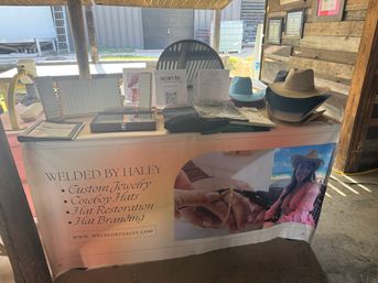 Rustic western market booth displaying custom jewelry tools, folded shirts and stacked cowboy hats with a promotional banner advertising custom jewelry, hat restoration and hat branding.