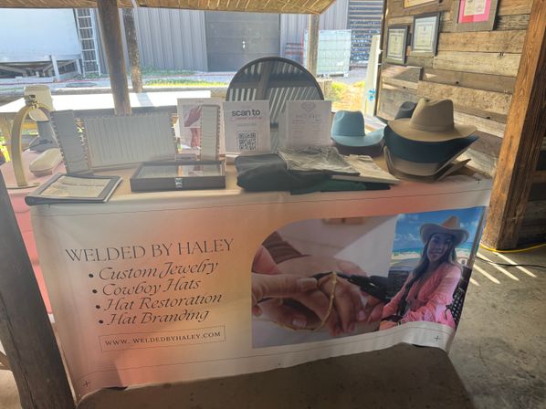 Rustic western market booth displaying custom jewelry tools, folded shirts and stacked cowboy hats with a promotional banner advertising custom jewelry, hat restoration and hat branding.