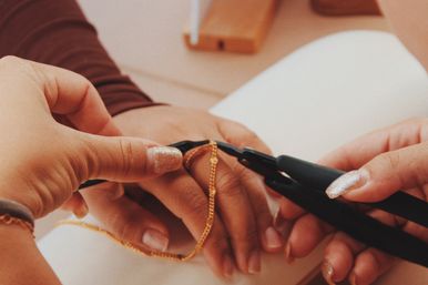 Close-up of hands with sparkly nails using pliers to fasten a delicate gold chain bracelet at a jewelry workbench