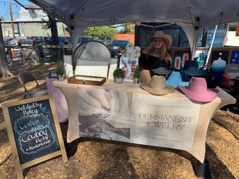 Outdoor craft market booth under a canopy displaying colorful cowboy hats, a permanent-jewelry banner and a chalkboard advertising custom hat restoration, with a vendor wearing a cowboy hat behind the table.