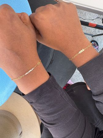 Close-up of two wrists wearing delicate personalized gold nameplate chain bracelets over a black long-sleeve, photographed outdoors on a concrete surface.