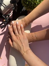 Sunlit close-up of hands with white manicure showcasing delicate gold bracelets and a pearl-accent hand chain on a pink tabletop
