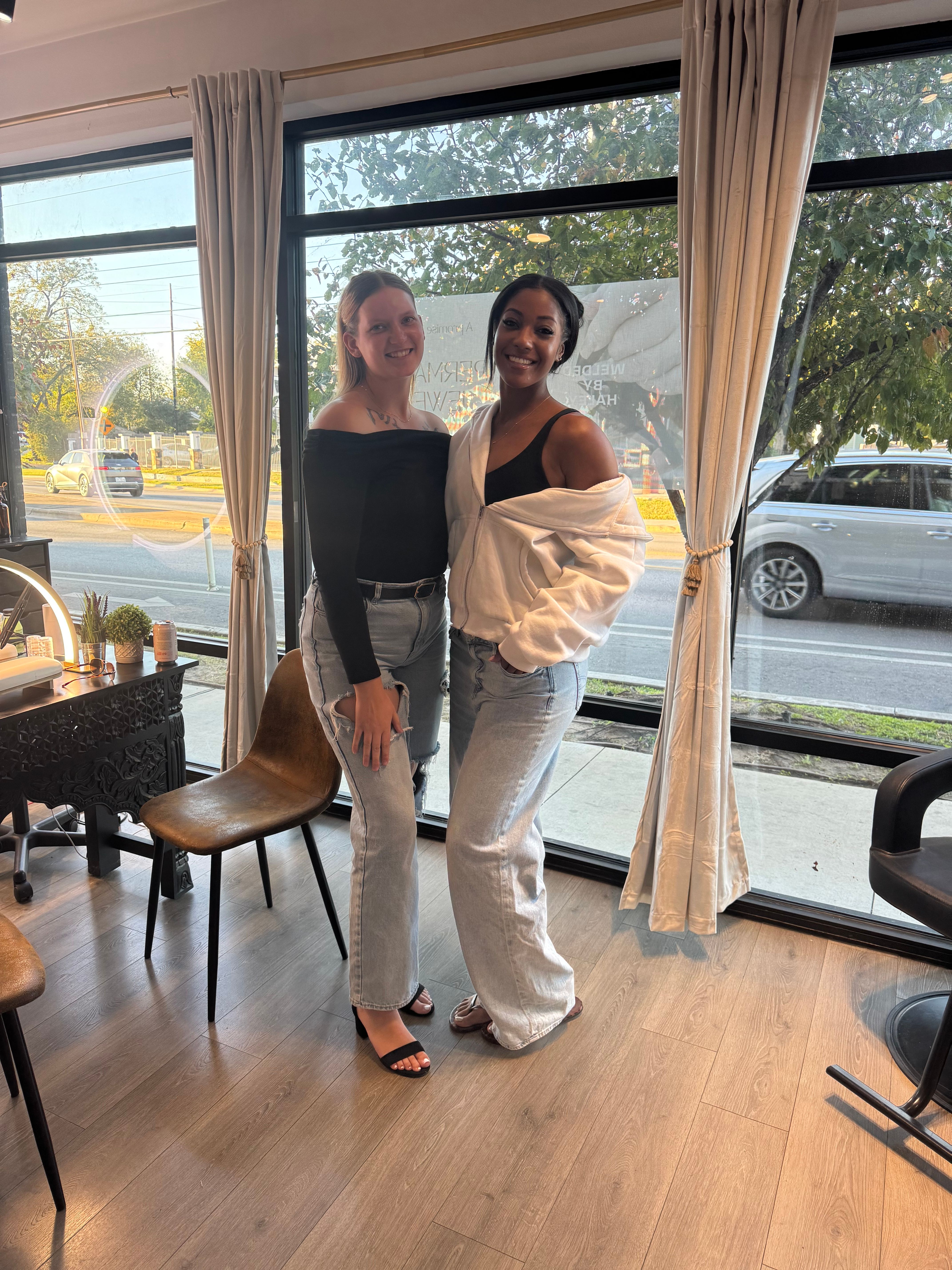 Two smiling friends posing inside a bright salon by floor-to-ceiling windows, wearing off-shoulder tops and light-wash jeans with a city street and cars visible outside.