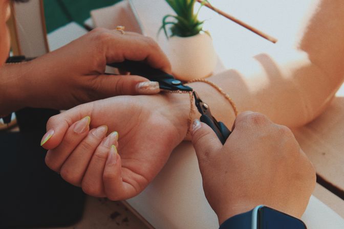 Close-up of hands using jewelry pliers to repair a delicate gold bracelet on a wrist, sunlit tabletop with a small potted succulent nearby