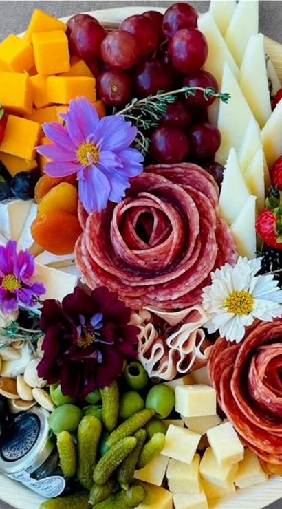 Vibrant round charcuterie board with salami roses, cheddar and assorted cheese cubes and wedges, red grapes, strawberries, blueberries and blackberries, dried apricots, mixed nuts, cornichons, green olives, and edible flowers on a wooden platter.