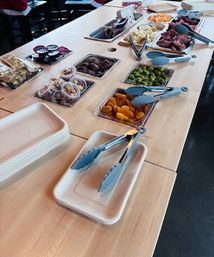 Self-serve charcuterie grazing station on a light wood table with compostable plates and tongs, trays of cured meats, cheeses, olives, dried apricots and mixed nuts.