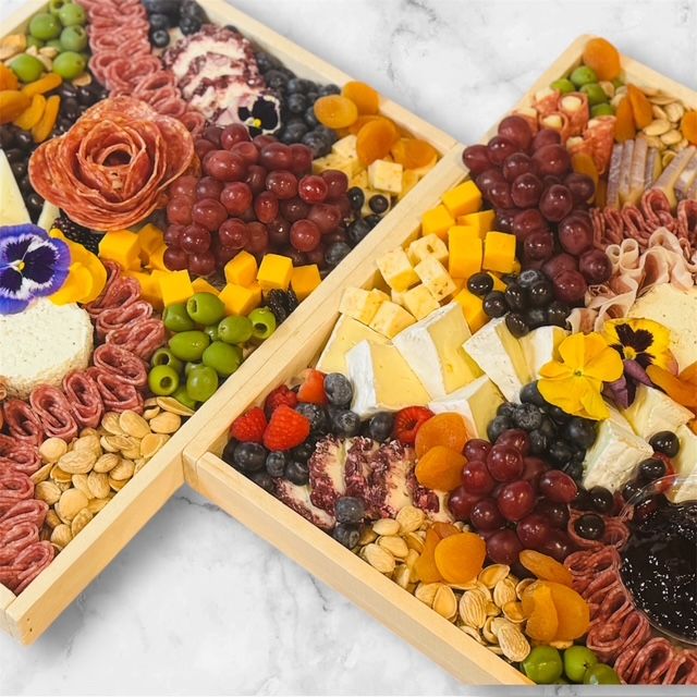 Gourmet wooden charcuterie boards on marble with brie wedges, cubed cheddar, salami roses, red grapes, green olives, dried apricots, mixed nuts, berries, jam and edible flowers — artisan cheese and meat party platter