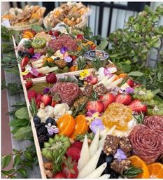 Colorful charcuterie board with salami rosettes, assorted cheeses, honeycomb, sliced oranges, strawberries, kiwi, berries, nuts, edible flowers and crackers on a wooden outdoor table with greenery