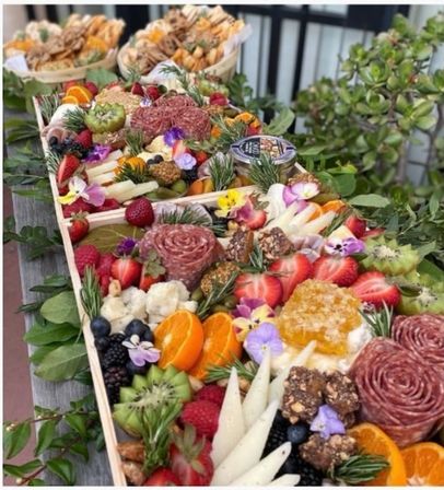 Colorful charcuterie board with salami rosettes, assorted cheeses, honeycomb, sliced oranges, strawberries, kiwi, berries, nuts, edible flowers and crackers on a wooden outdoor table with greenery