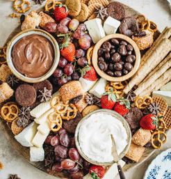 Dessert charcuterie grazing board with bowls of chocolate and whipped cream dips, chocolate-covered nuts, strawberries, red grapes, assorted cookies, wafer sticks, pretzels and sliced cheese on a round platter