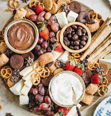 Dessert charcuterie grazing board with bowls of chocolate and whipped cream dips, chocolate-covered nuts, strawberries, red grapes, assorted cookies, wafer sticks, pretzels and sliced cheese on a round platter