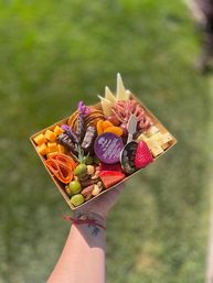 Colorful handheld charcuterie box for an outdoor picnic — cubed and sliced cheeses, folded cured meats, crackers, strawberries, olives, mixed nuts, dried apricots and lavender garnish over green grass