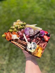 Handheld charcuterie grazing box over grass with cubed cheddar and sliced cheeses, rolled cured meats, green olives, red grapes, strawberries and blueberries, crisp crackers, chocolate bites and a wooden fork