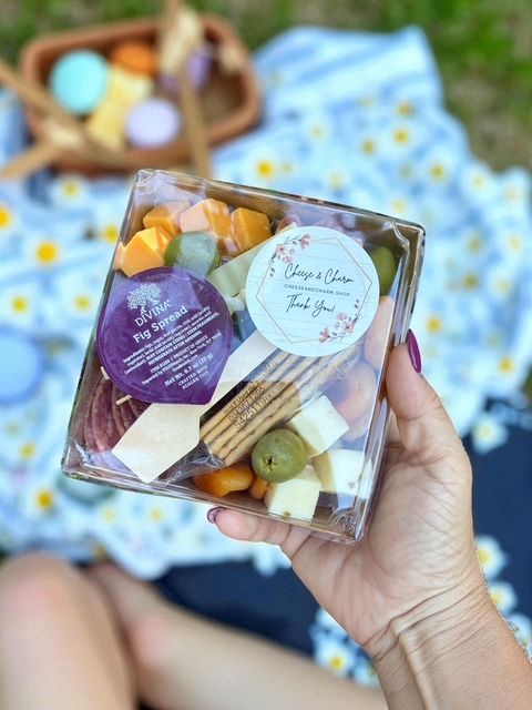 Hand holding a clear plastic picnic snack box with cubed cheeses, crackers, green olive and a small jam cup, set over a daisy-print picnic blanket in a sunny outdoor park.