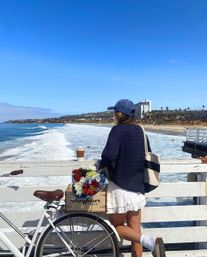 Person in a navy sweater and baseball cap leans on a pier railing next to a bicycle with a crate of colorful flowers, overlooking bright blue ocean waves, sandy beach and palm-lined coastal skyline under a clear sky.