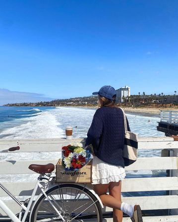 Person in a navy sweater and baseball cap leans on a pier railing next to a bicycle with a crate of colorful flowers, overlooking bright blue ocean waves, sandy beach and palm-lined coastal skyline under a clear sky.