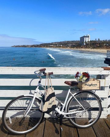 White vintage bicycle with a striped tote and crate of flowers leaned against a pier railing, overlooking surfers on small waves and a sandy coastal beach with bluffs and a tall oceanfront building under a bright blue sky.