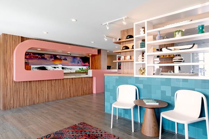Coastal-inspired boutique hotel lobby with pastel pink curved reception desk, sunset ocean mural, teal-tiled front, white waiting chairs, small wooden table and decorative shelving.